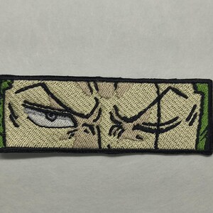 May include: Embroidered patch featuring a stern, stylized face with a single visible eye. The design uses black and tan thread, with green borders. The patch is rectangular and likely intended for application on clothing or accessories.