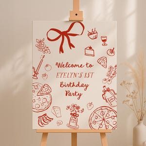 May include: A cream-coloured sign on a wooden easel, featuring red illustrations of food and a bow. The sign reads "Welcome to Evelyn's 1st Birthday Party."