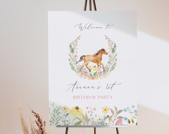 May include: Welcome to Ariana's 1st Birthday Party sign featuring a watercolour illustration of a brown horse running through a wreath of flowers. The sign is on a black easel.