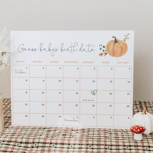 Peut inclure: Un calendrier blanc avec un graphique de citrouille et le texte "Devinez la date de naissance du bébé". Le calendrier affiche les mois d'octobre et de novembre avec la date d'échéance marquée le 19 octobre.