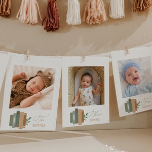 May include: Three white photo frames with brown wooden clips hanging on a wall with a garland of brown wooden beads and tassels. The frames feature photos of a baby at different ages, with the text "Two Months", "Three Months", and "Four Months" on each frame.