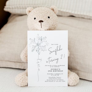 May include: A white invitation card featuring a silver snowflake design. The text reads "Our Little Snowflake is turning 2!" and includes details for Everly's 2nd birthday party. The card is held by a white teddy bear.