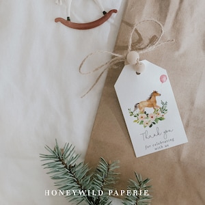 Puede incluir: Una bolsa de regalo de papel marrón con una etiqueta blanca atada con una cuerda. La etiqueta presenta una ilustración de acuarela de un caballo marrón con un globo rosa, rodeado de flores. El texto en la etiqueta dice "Gracias por celebrar con nosotros".