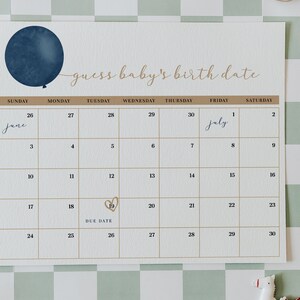 Op de afbeelding: Een beige kalender met de tekst "guess baby's birth date" en een blauwe ballongrafiek. De kalendergrid is beige met goudkleurige lijnen en data. De vervaldatum is gemarkeerd op de 19e van de maand. De woorden "June" en "July" zijn geschreven.