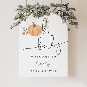 Peut inclure: Une pancarte blanche avec le texte "It's a baby" en écriture cursive noire, une illustration aquarelle d'une citrouille et le texte "Welcome to Everly's Baby Shower". La pancarte est suspendue à une branche de feuilles d'eucalyptus.