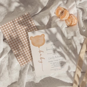 May include: A gender reveal invitation with a bear balloon illustration and the text "He or She, we can bearly wait." The invitation is on a cream background with a gingham patterned card and a peeled tangerine.
