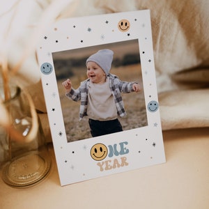 May include: A white card with a photo of a child in a blue and white plaid shirt. The photo is surrounded by a white frame with blue and yellow smiley faces and stars. The text "ONE YEAR" is written in blue and yellow letters with a smiley face.