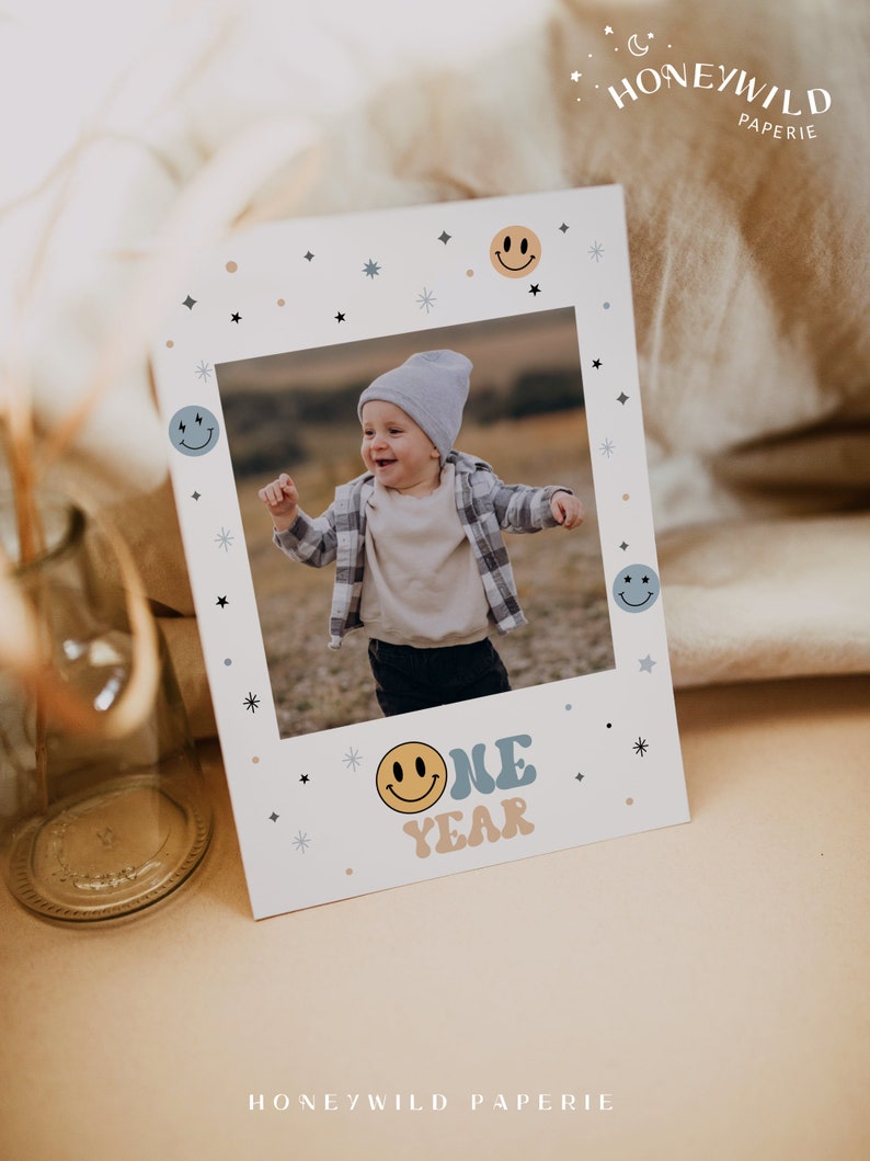 May include: A white card with a photo of a child in a blue and white plaid shirt. The photo is surrounded by a white frame with blue and yellow smiley faces and stars. The text "ONE YEAR" is written in blue and yellow letters with a smiley face.