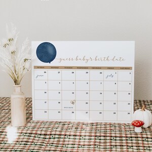Op de afbeelding: Een witte kalender met een marineblauwe ballongrafiek en de tekst "guess baby's birth date". De kalender markeert de vervaldatum van 19 juni met een gouden hart. Een vaas met gedroogde bloemen en decoratieve items staan op de voorgrond.