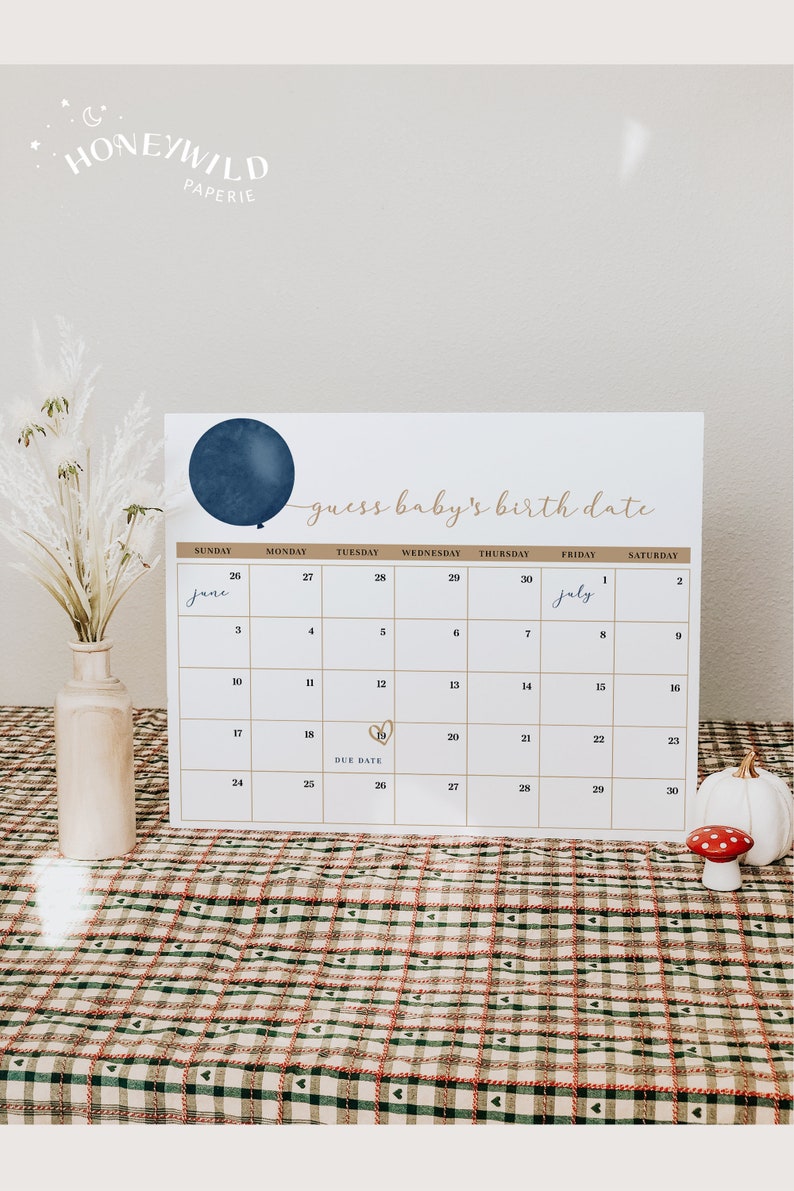 Op de afbeelding: Een witte kalender met een marineblauwe ballongrafiek en de tekst "guess baby's birth date". De kalender markeert de vervaldatum van 19 juni met een gouden hart. Een vaas met gedroogde bloemen en decoratieve items staan op de voorgrond.