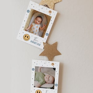 May include: A set of two white photo cards with a smiling face and the text "Three Months" and "Four Months" on each card. The cards are decorated with blue and yellow smiley faces and stars. The cards are hanging from a string with gold glitter stars.