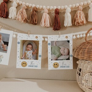 May include: Three white photo frames with a smiling face icon and the text "Three Months" and "Four Months". The frames are hanging on a wall with a white and brown tassel garland above them.