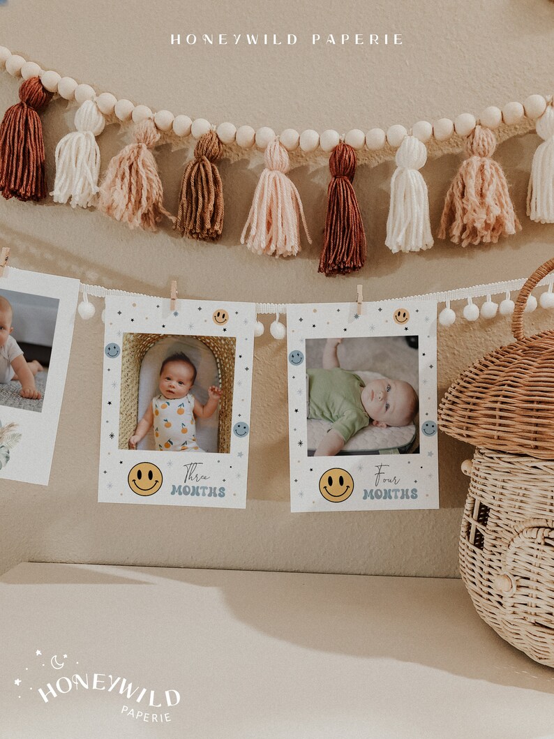 May include: Three white photo frames with a smiling face icon and the text "Three Months" and "Four Months". The frames are hanging on a wall with a white and brown tassel garland above them.