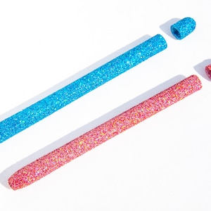 May include: Two glitter-covered makeup applicators, one blue and one pink, with the caps removed.