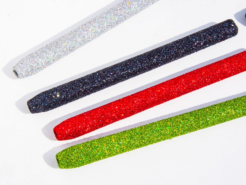 May include: Four glitter-covered pens in different colors: silver, black, red, and green. The pens are arranged in a diagonal line on a white surface.