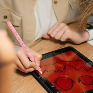 May include: A person uses a pink glitter stylus to draw a red and black cartoon face on a digital tablet.