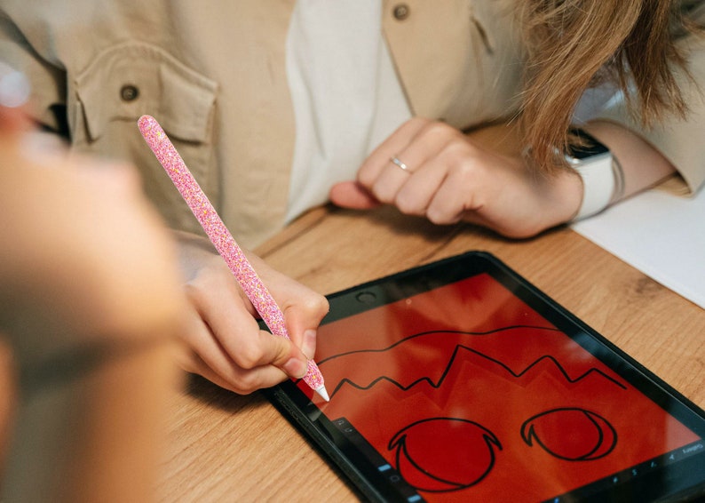 May include: A person uses a pink glitter stylus to draw a red and black cartoon face on a digital tablet.