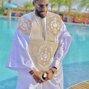 May include: A man wearing a white and gold embroidered robe with a gold watch on his wrist. He is standing in front of a swimming pool.
