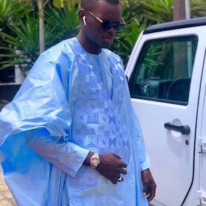 May include: A man wearing a light blue traditional robe with a geometric pattern, sunglasses, and a gold watch. He is standing next to a white car.