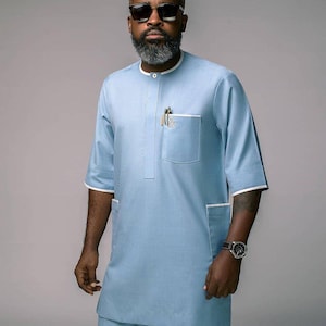 May include: Light blue tunic and pants set with white trim. The tunic has a front pocket and a mandarin collar. The outfit is worn with sunglasses and a wristwatch.