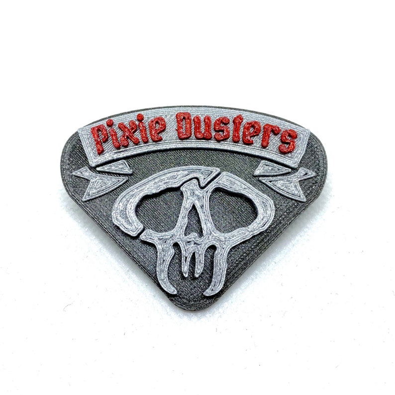 Puede incluir: Un pin de metal plateado con un dise&ntilde;o de calavera y el texto "Pixie Dustlers" en letras rojas.