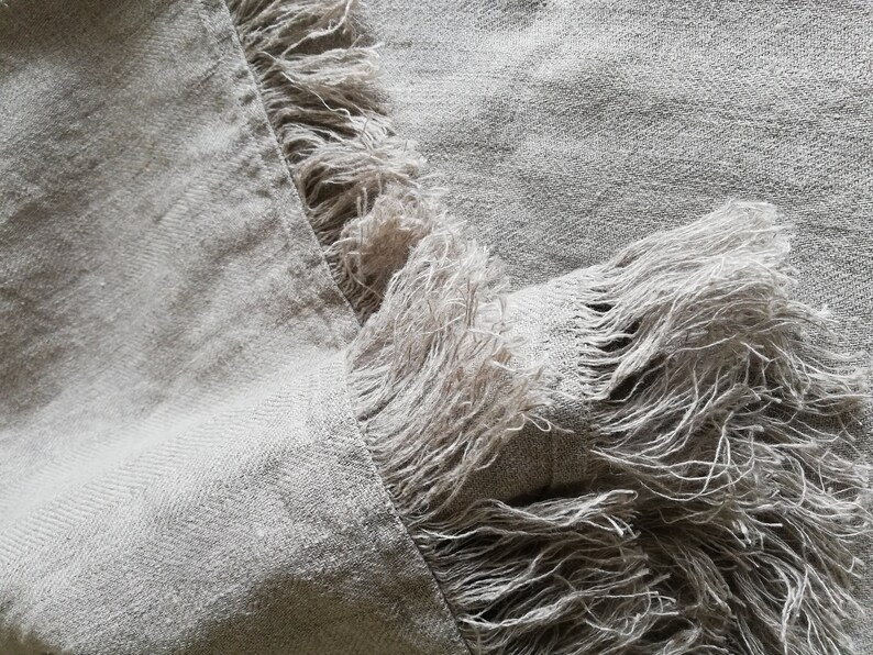 May include: A close-up of a light gray linen fabric with a fringe border. The fabric has a subtle herringbone weave.