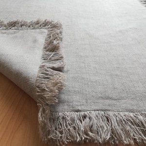May include: A light grey linen rug with a fringed border. The rug is folded in half and the fringe is visible on both sides.