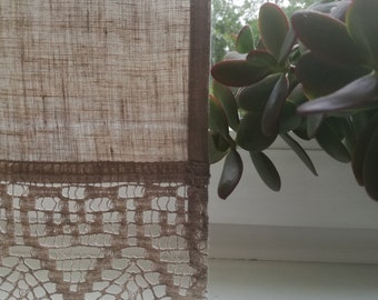 May include: A beige linen curtain with a lace trim hangs in a window. A green plant with red leaves is visible outside the window.