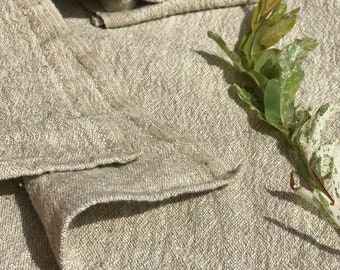 May include: A stack of folded linen towels in a natural beige color. The towels are folded and arranged on a textured surface with a sprig of green leaves.