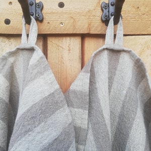 May include: Two gray and white striped linen tea towels hanging on black hooks attached to a wooden wall.