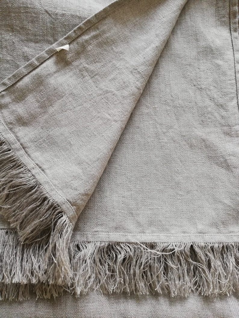 May include: A light gray linen throw blanket with a fringe border. The blanket is folded in half and the fringe is visible on both sides.