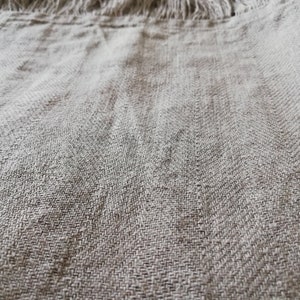 May include: Close-up of a light brown linen fabric with a frayed edge. The fabric has a woven texture and a subtle sheen.