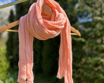 May include: A peach-colored scarf hangs from a wooden hanger on a metal bar. The scarf has a textured, pleated design and is made of a lightweight fabric. The background features green trees and a blue sky, suggesting an outdoor setting.