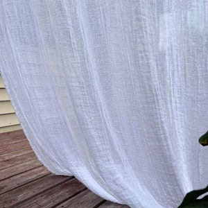 Può includere: Pannello di tenda bianca trasparente con un aspetto testurizzato e stropicciato. La tenda è appesa su un ponte di legno.