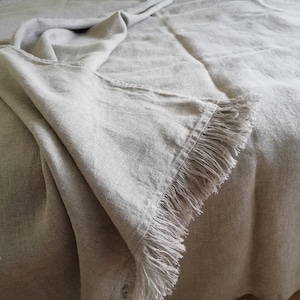 May include: A light gray linen throw blanket with a fringe border. The blanket is folded and draped over a bed.