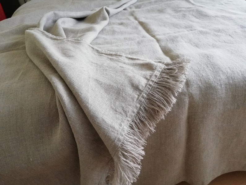 May include: A light gray linen throw blanket with a fringe border. The blanket is folded and draped over a bed.