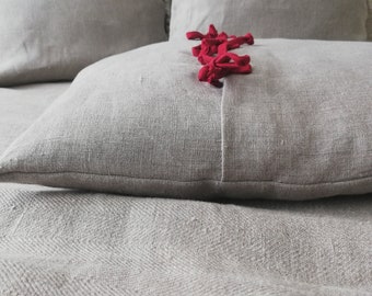 May include: A close-up of a gray linen pillow with a red ribbon tied in a knot in the center. The pillow is on top of a gray linen bedspread.