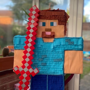 May include: A Minecraft-themed piñata of a character holding a red and white sword. The character has a tan face, blue eyes, and a brown beard and hair. The shirt is blue, and the trousers are blue and black.
