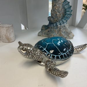 May include: A decorative turtle figurine with a silver-colored body and a blue-green shell. The turtle is lying on a white surface. The shell has a glossy finish. The figurine is a decorative item.