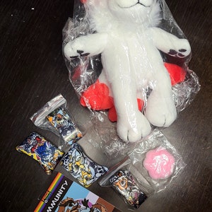 May include: A white and red plush toy of a fox-like creature in a plastic bag. There are five small bags with colorful printed fabric inside. One bag contains a pink flower-shaped object. A card with the text "Pride Pins Community is about unity" is on a wooden surface.