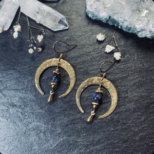 May include: A pair of gold crescent moon earrings with blue gemstone beads hanging from the bottom. The earrings have a hammered texture and are accented with small decorative circles.