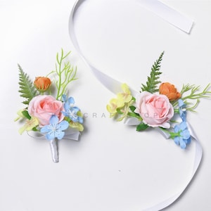 Op de afbeelding: Twee bloemenpols corsages met witte linten. Elke corsage heeft een roze roos, een oranje knop, blauwe en gele accentbloemen en groen blad. De corsages zijn waarschijnlijk voor een bruiloft of een speciale gelegenheid.