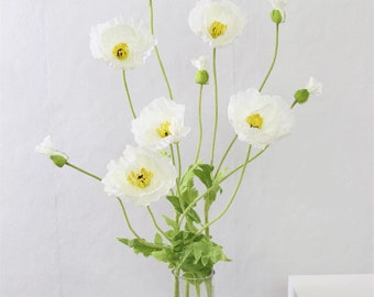 Puede incluir: Un ramo de flores de amapola blancas con centros amarillos en un jarrón de vidrio transparente. Las flores están dispuestas en un estilo simple y elegante.
