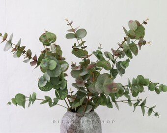 May include: Eucalyptus branches with round, green leaves and hints of purple and red, arranged in a textured gray vase. The image features the text "RITA CRAFTSHOP" at the bottom of the vase.