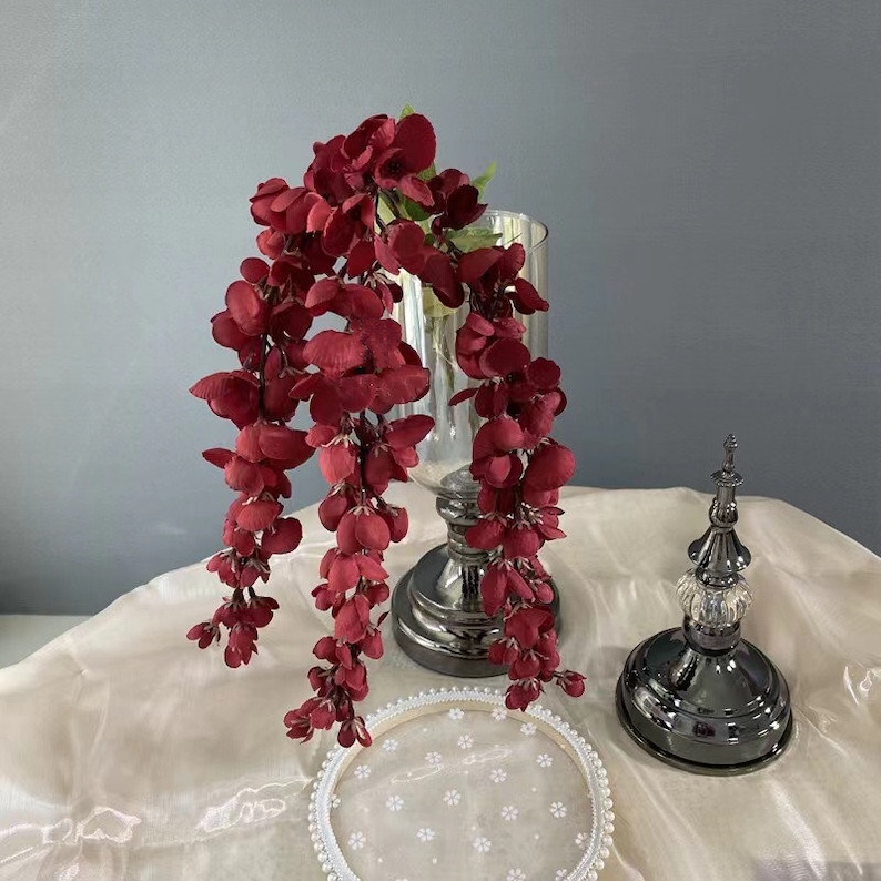 Pu&ograve; includere: Un vaso di vetro con base argentata e una cascata di fiori artificiali rosso scuro. Il vaso &egrave; appoggiato su una tovaglia bianca con un pizzo bianco.