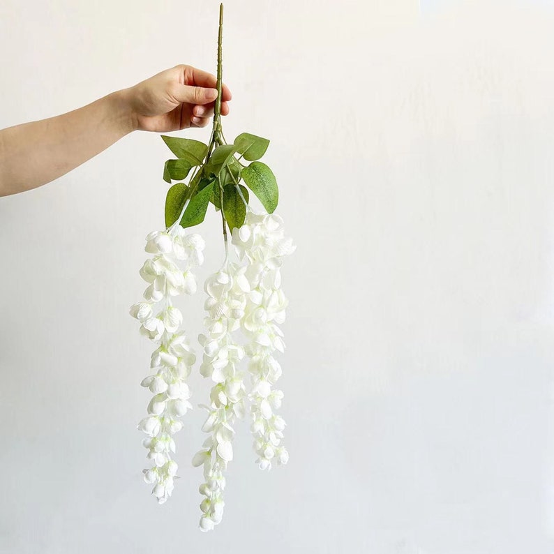 Pu&ograve; includere: Una ghirlanda di fiori di glicine artificiali bianchi con foglie verdi. La ghirlanda pende a cascata di fiori.