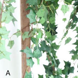 May include: A close-up of a green artificial ivy vine with a brown wooden stick in the center. The vine is hanging down and the leaves are in focus.