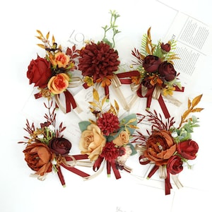 Könnte beinhalten: Sechs künstliche Blumen-Boutonnières in Rot-, Orange- und Brauntönen. Jede Boutonnière besteht aus verschiedenen Blumen, darunter Rosen, Pfingstrosen und Nelken, und ist mit Grün und einem burgunderfarbenen Band verziert.