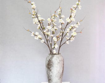 Puede incluir: Un ramo de ramas de cerezo artificial blanco en un jarrón de cerámica gris y blanco. El jarrón está sobre un pedestal blanco.