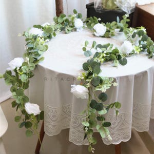 以下が含まれることがあります： 白い人工の花と緑色のユーカリの葉のガーランドが、レースの縁取りのある白いテーブルにかけられています。ガーランドは、結婚式やパーティーの飾り付けに最適です。